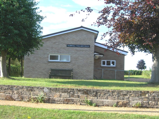 vh1 Small brick building labeled Yarwell Village Hall with a bench and trees in front.