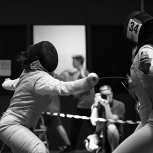 Two fencers in action during a match, with people and a photographer in the background.