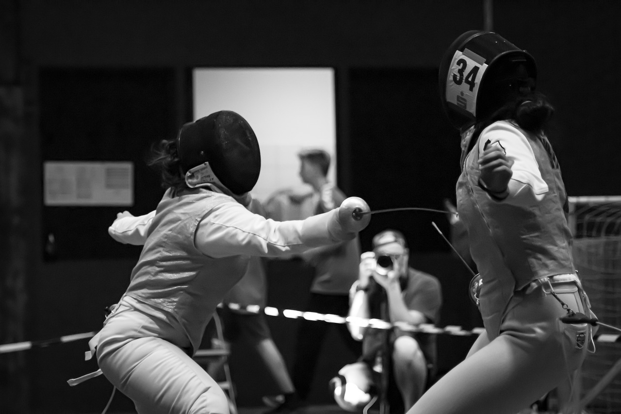 Two fencers in action during a match, with people and a photographer in the background.