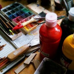 Colorful art supplies: colored pencils, paint bottles, watercolors, and brushes on a messy table.