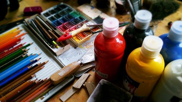 Colorful art supplies: colored pencils, paint bottles, watercolors, and brushes on a messy table.